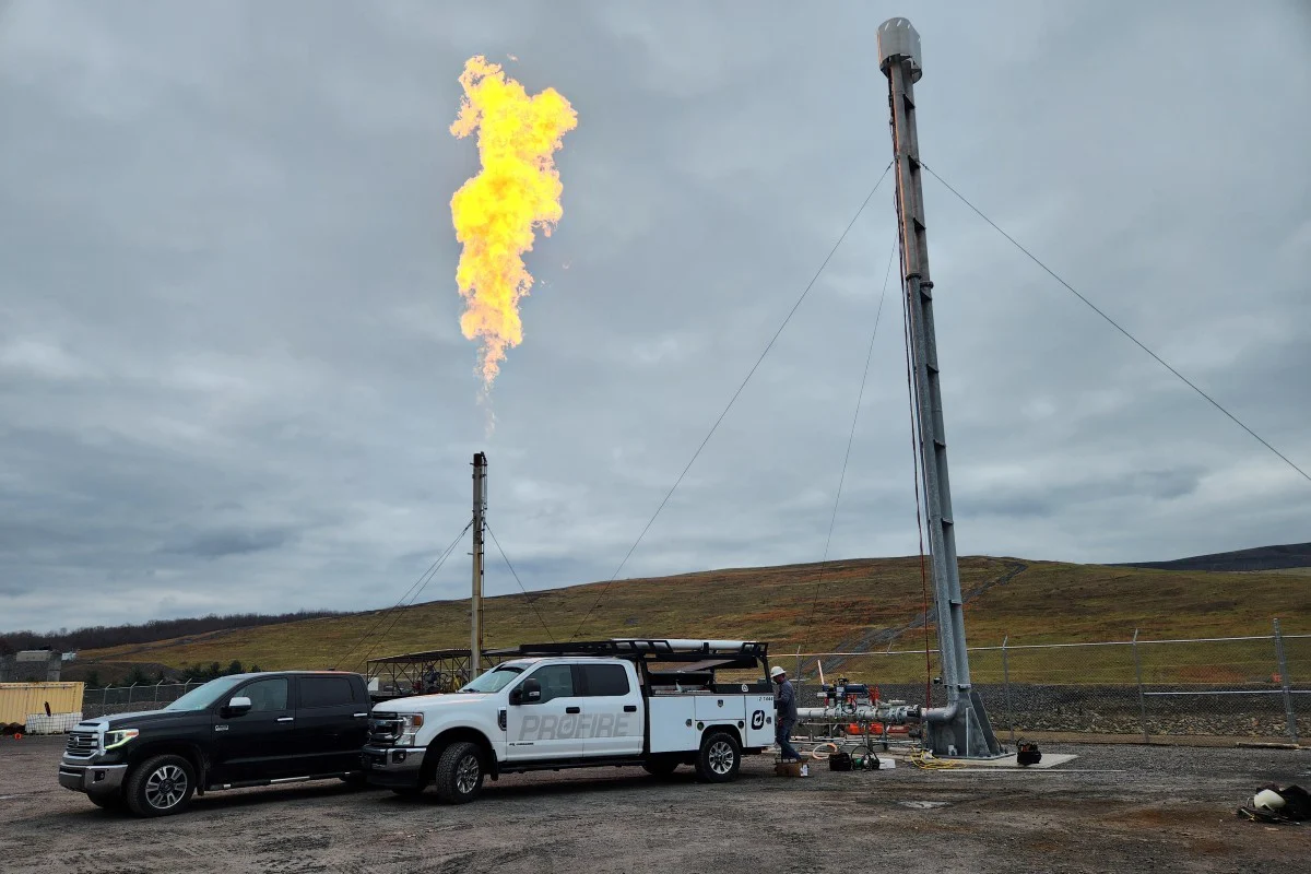 The photo depicts two flare systems at a RNG landfill location. One on the right is taller and is a dual candlestick flare. The left is a shorter flare emitting a flame. In the foreground there are two trucks, one black and one white with Profire decals on it.