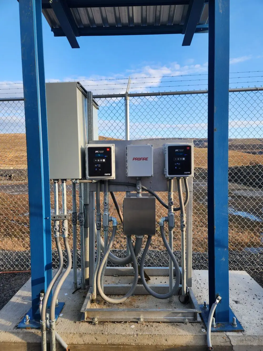The picture shows a concrete pad with a BMS controller system sitting atop. There are two controllers linked by a central communications unit. There's a mechanical box to the left of the controllers which sit in the middle of the image. Above there's a tin roof providing cover for these systems. Behind is a field with hills and a blue sky.