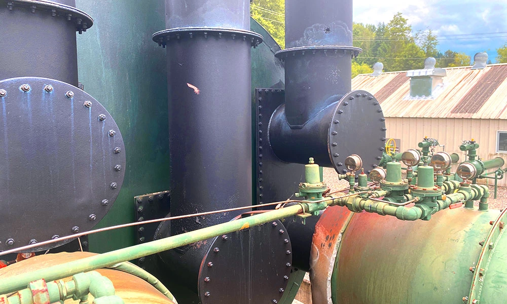 Close-up of black exhaust/duct piping and a green valve manifold with pressure gauges; visible corrosion on the vessel surface.