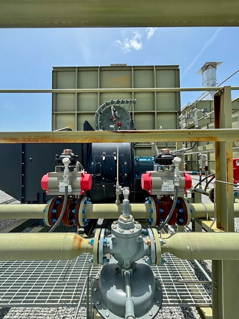 Close-up of a gas train with flanged piping, regulator, instrumentation, and two red actuator-controlled valves mounted in front of a large industrial burner assembly.