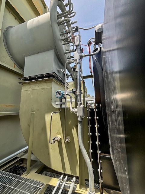 Side view of an industrial burner or air register assembly with curved piping, instrumentation, and conduit mounted alongside a large cylindrical vessel.