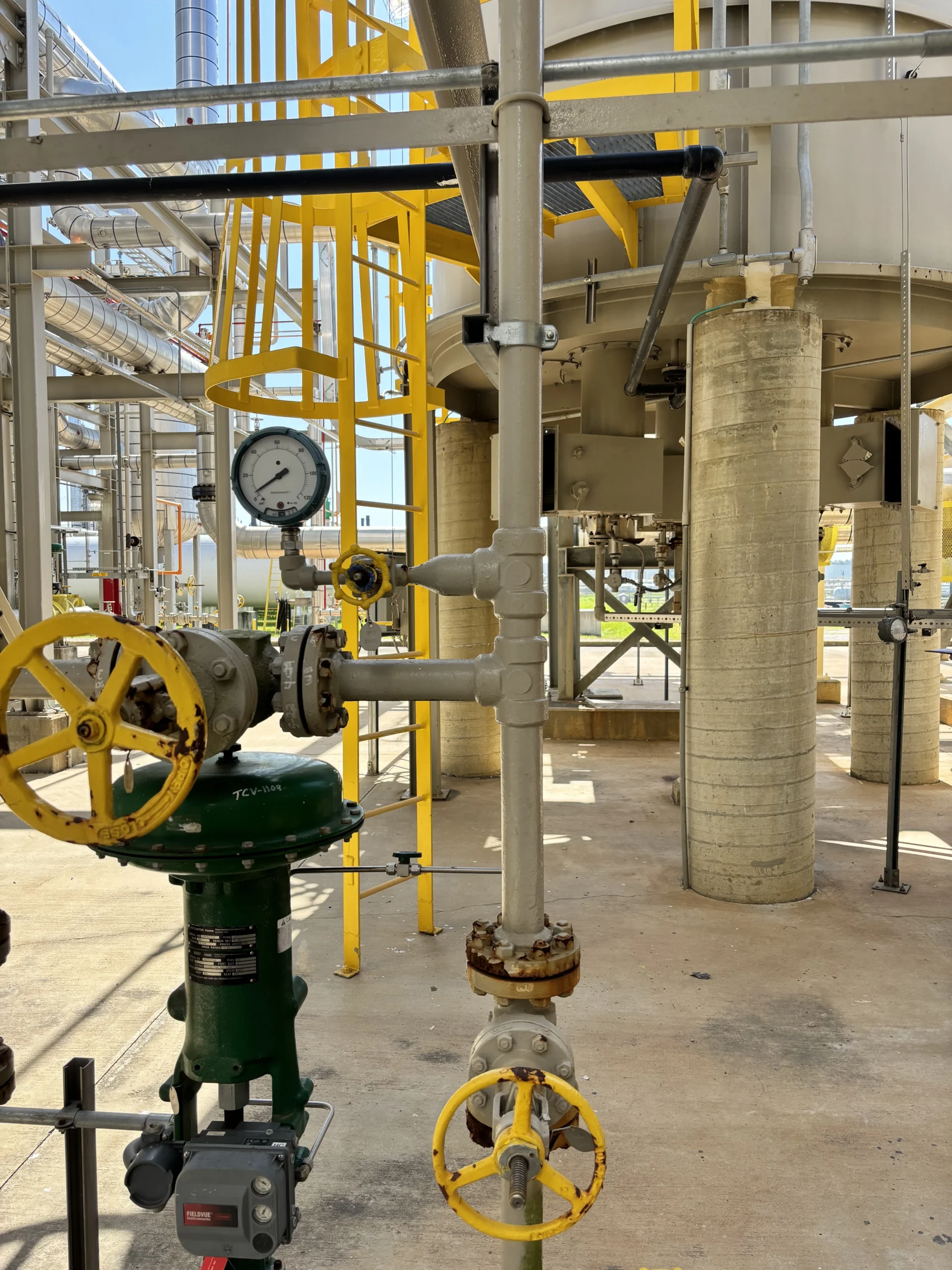 Close-up of fuel gas piping, a yellow handwheel valve, pressure gauge, and green valve actuator beneath an industrial process heater.