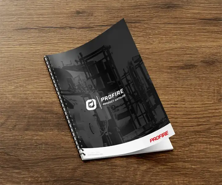 Spiral-bound Profire product catalog booklet with dark industrial cover photo, resting at an angle on a wooden desk surface.