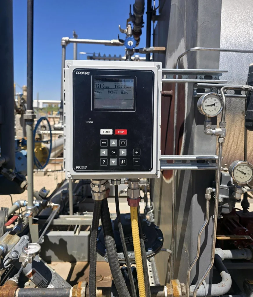 Industrial outdoor setup featuring a Profire PF2200-GP controller panel mounted on piping; the rectangular control unit has a digital display showing readings, “START” and “STOP” buttons, and a keypad. Surrounding the panel are metal pipes, valves, and pressure gauges attached to a large cylindrical vessel, with cables running from the bottom of the controller. The equipment is installed in an open facility under a clear blue sky.