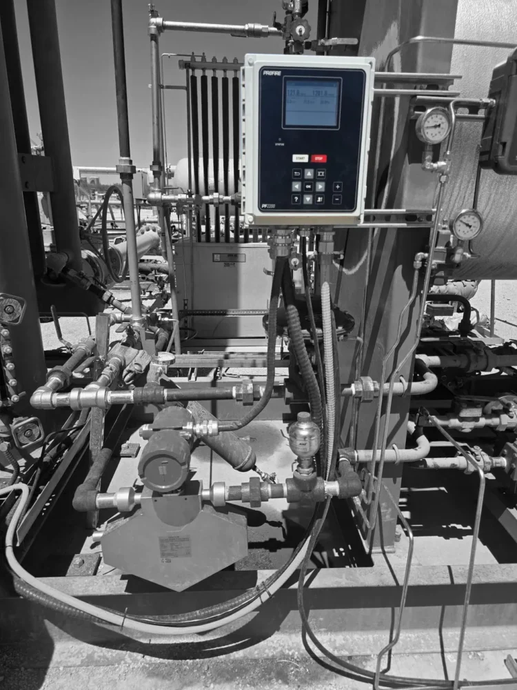 Black-and-white image of an industrial process setup featuring a Profire Controller mounted on metal piping. The rectangular control panel has a digital display and keypad with labeled buttons, connected by multiple cables running downward. Surrounding the controller is a network of pipes, valves, and fittings, along with pressure gauges mounted on a large cylindrical tank to the right. Additional equipment, including a pump and tubing, is visible in the foreground, indicating an outdoor industrial environment such as a gas or process facility.