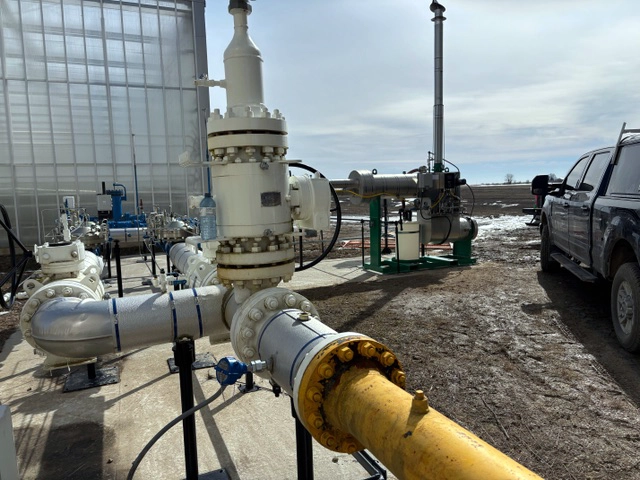 Wide view of a gas pipeline and valve assembly connected to a skid-mounted fired heater beside a greenhouse.