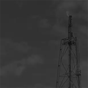 Dark, low-contrast photo of a tall oilfield flare stack silhouetted against a cloudy sky.