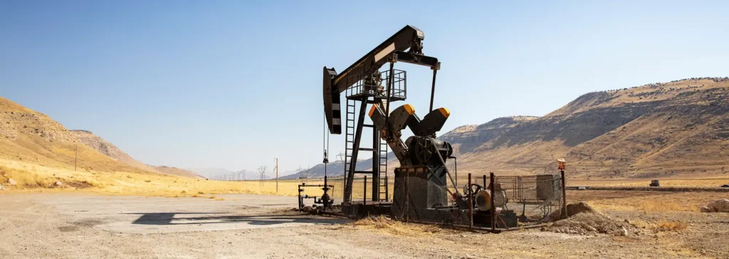 An oil pump jack on a bare dirt pad in an arid landscape of dry golden hills, with power lines in the distance under a clear blue sky.