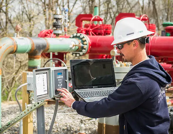 A technician in a white Profire hard hat and sunglasses uses a laptop and a control panel at a wellsite, with red and green pipes behind.