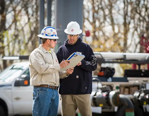 Two workers in hard hats look at a clipboard at an industrial oilfield site, with a white pickup truck and horizontal piping behind them.