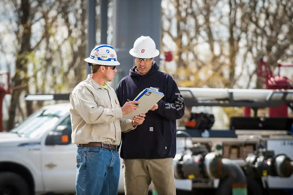 Two workers in hard hats review a clipboard at a wellsite, with a white pickup truck and piping behind them.