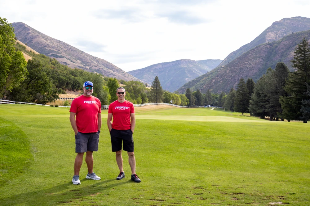 Two people wearing red PROFIRE shirts standing on a golf course surrounded by trees and mountains during the 2026 Burn Camp golf tournament.